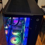 Black PC tower with a tempered-glass side panel showing RGB-lit fans and cables inside. The top has a vented grill and a power button on the front panel.