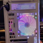 White mid-tower PC case with perforated side panels glowing purple and blue RGB, front I/O ports visible (USB, audio).“