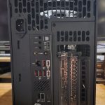 Rear view of a black desktop PC showing the motherboard I/O panel, power inlet, and multiple PCIe expansion slots behind a vented panel; a cooling fan is visible through the grill.