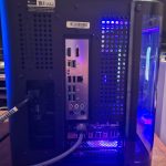 Rear view of a desktop PC tower with blue LED lighting, showing the I/O panel with USB and video ports and perforated ventilation.